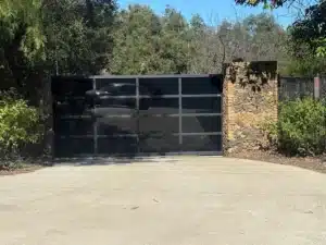 A wide driveway gate featuring dark tinted glass panels in a black metal frame, set against a rustic stone wall.