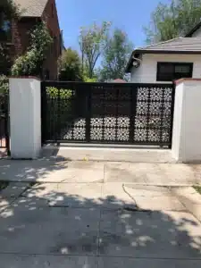 A black sliding metal driveway gate with a decorative geometric circular pattern between two white concrete pillars.