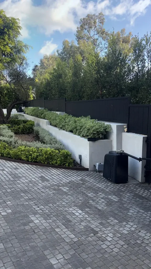 A white tiered retaining wall with manicured shrubs alongside a black privacy fence and cobblestone motor court.