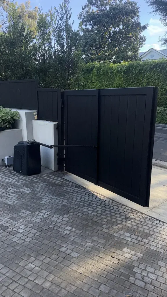 sleek, matte black privacy gate standing open over a cobblestone driveway, showcasing the automated arm and clean white concrete pillars.