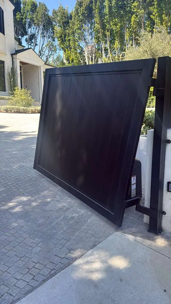 Automated black steel barn-style gate partially open, revealing a luxury residential driveway and modern home exterior in the background.