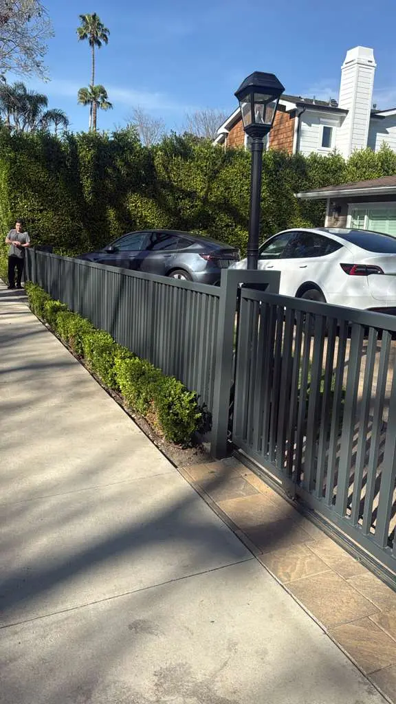 Low angle view of a dark gray vertical slat metal fence running parallel to a sidewalk with small green bushes at the base.