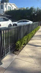 A perspective view showing a long stretch of modern gray steel fencing along a sidewalk, securing a residential property with parked cars visible inside.