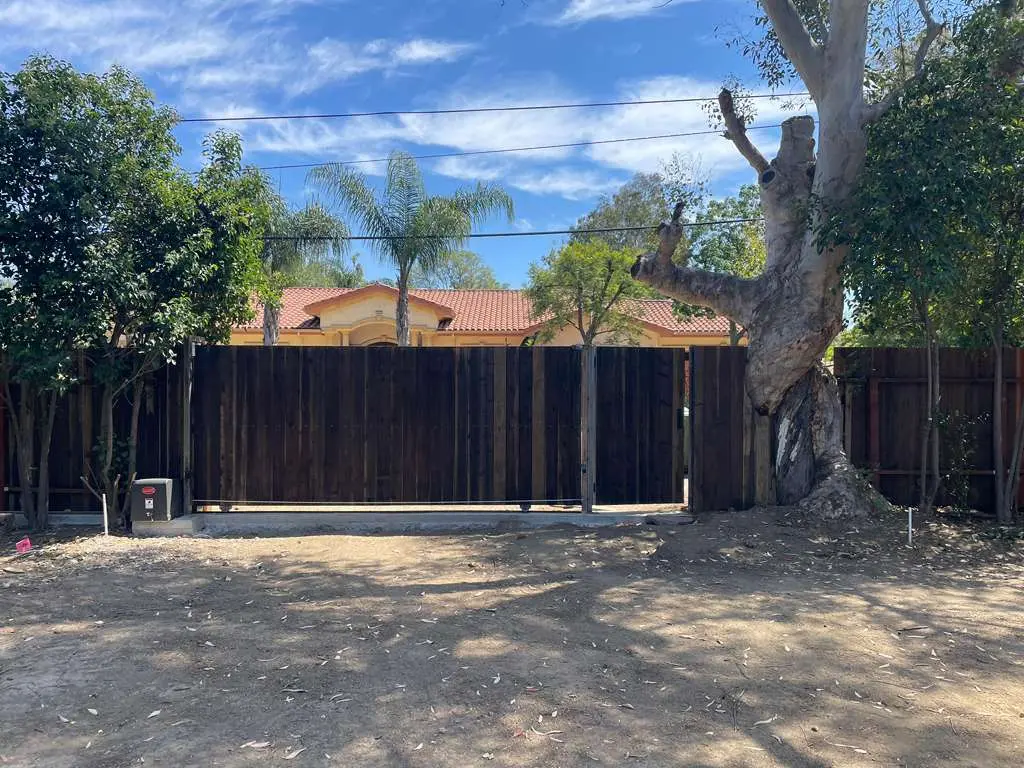 A wide view of a continuous dark vertical wood plank fence bordering a property line, flanked by mature trees and a dirt foreground.