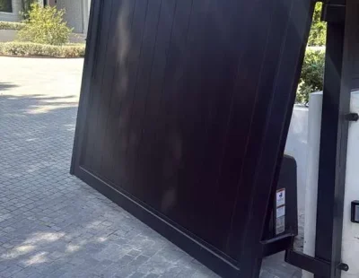 Automated black steel barn-style gate partially open, revealing a luxury residential driveway and modern home exterior in the background.