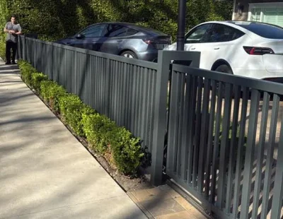 Low angle view of a dark gray vertical slat metal fence running parallel to a sidewalk with small green bushes at the base.