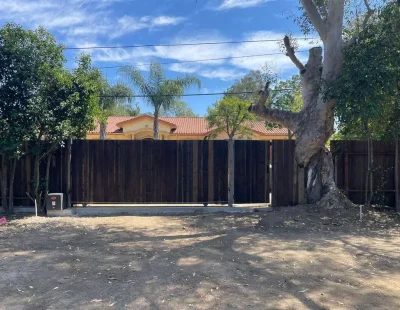 A wide view of a continuous dark vertical wood plank fence bordering a property line, flanked by mature trees and a dirt foreground.